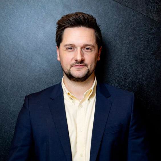 A man with short dark hair stands against a dark background. He is wearing a navy blue blazer and a light yellow shirt. He looks friendly at the camera and has a well-groomed beard