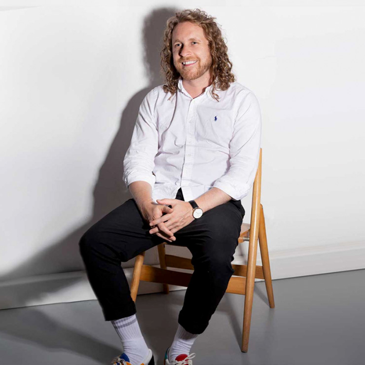 A smiling man with curly hair is sitting on a wooden chair in front of a white background. He is wearing a white shirt, black pants, and colorful sneakers.